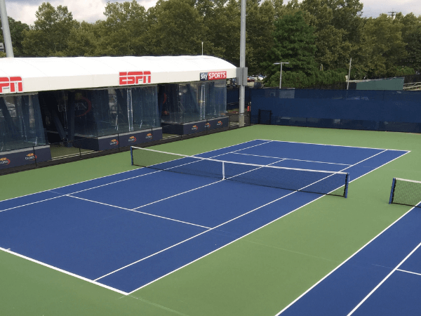 Blue and green outdoor tennis court with ESPN and Sky Sports broadcast booths in the background, surrounded by trees and fencing.