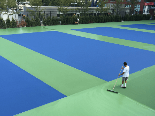 Person using a roller to apply green paint on an outdoor sports court with several blue rectangular sections.