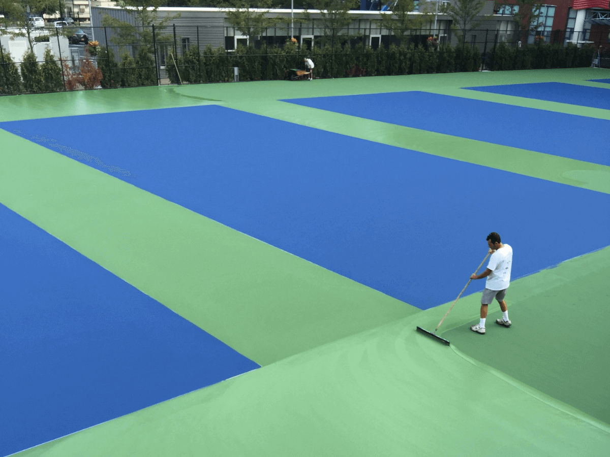 A person uses a large squeegee to spread green paint or surface material on an outdoor tennis court with blue sections.