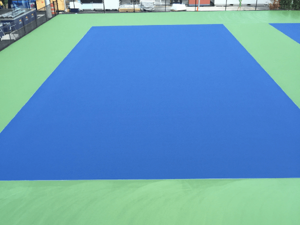 A blue rectangular tennis court with a green outer area, viewed from above.