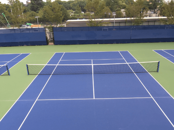 An empty outdoor blue and green tennis court with a net in the center, surrounded by fencing and trees in the background.