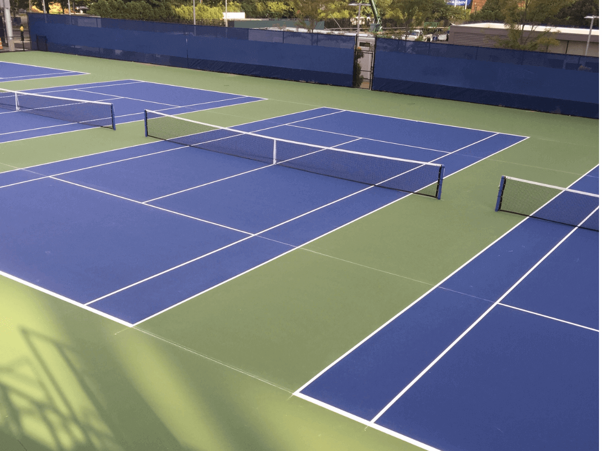 Several empty blue and green hard tennis courts are lined up outdoors, separated by nets and surrounded by a blue fence.