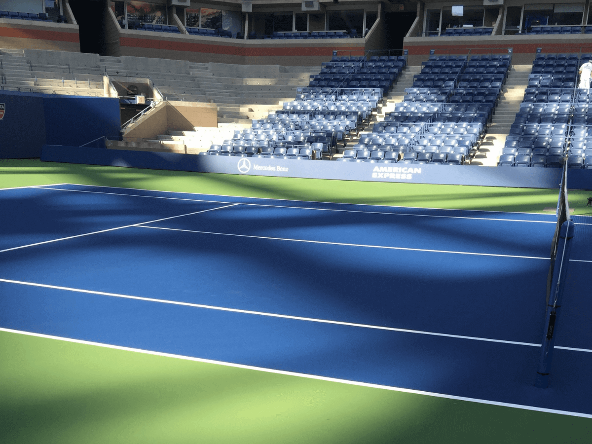 Empty blue and green tennis court inside a stadium with blue seating and sponsor logos visible on the stands.