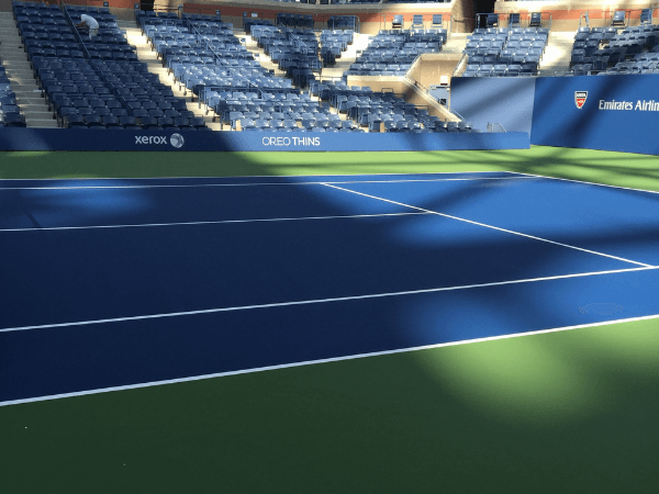 A blue and green outdoor tennis court with empty blue stadium seats and sponsorship banners, partially shaded by sunlight.