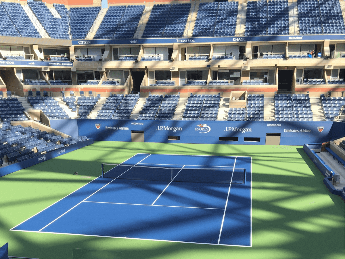 Empty tennis court at Arthur Ashe Stadium, with blue seats and sunlight casting shadows across the court and stands.