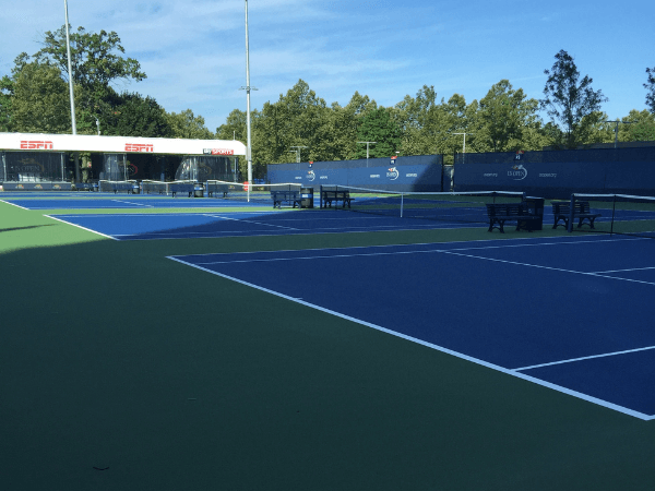 Multiple outdoor tennis courts with blue playing surfaces and green borders, benches along the sidelines, and an ESPN broadcast booth in the background.