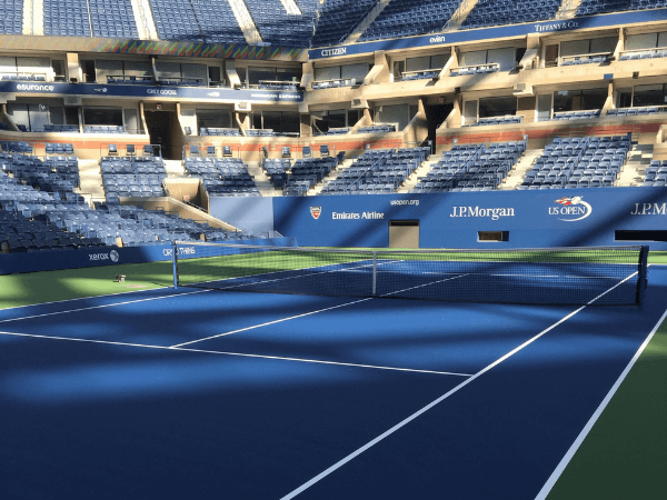 Empty tennis court with blue surface and white lines inside a large stadium with rows of blue seats and sponsor banners.