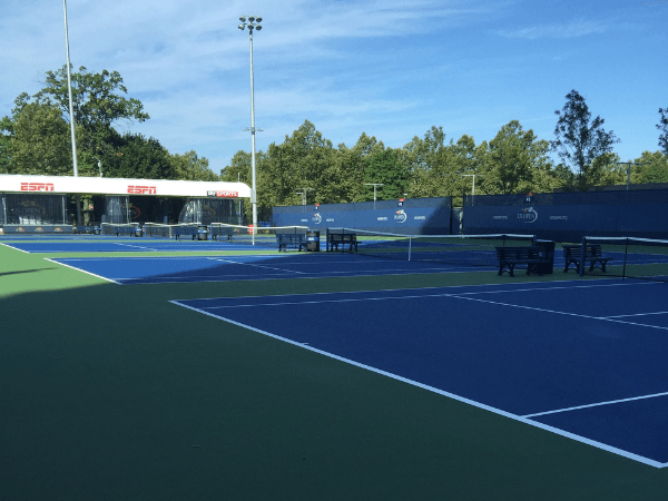 Multiple empty blue and green outdoor tennis courts with benches and an ESPN broadcast booth in the background. Trees and blue sky are visible.