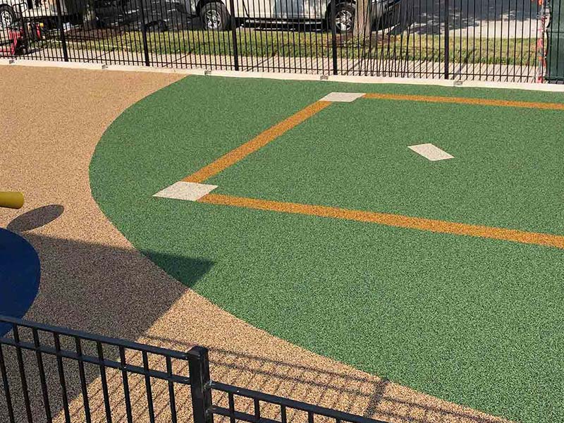 A small fenced-in playground area featuring a green and tan rubber surface designed to resemble a baseball diamond.