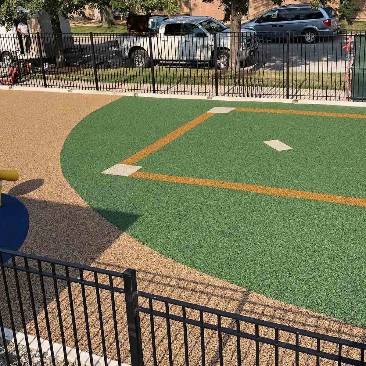 View of a small, fenced playground area with a green and tan rubber surface marked to resemble a baseball field; vehicles are parked on the street outside the fence.