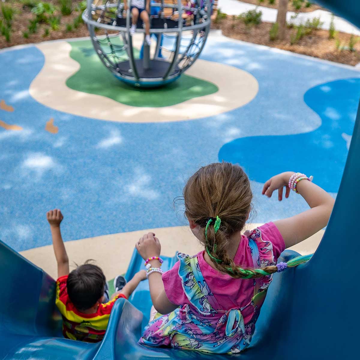 Two children slide down a blue playground slide while another child climbs inside a spherical climbing structure in the background.