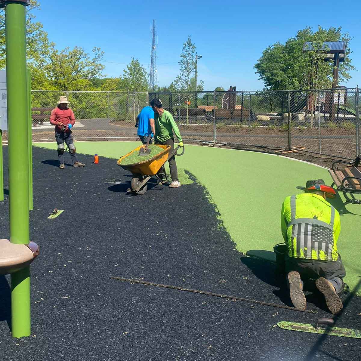 Three workers install or repair rubber surfacing on a playground, using a wheelbarrow and tools, with fencing and trees in the background.