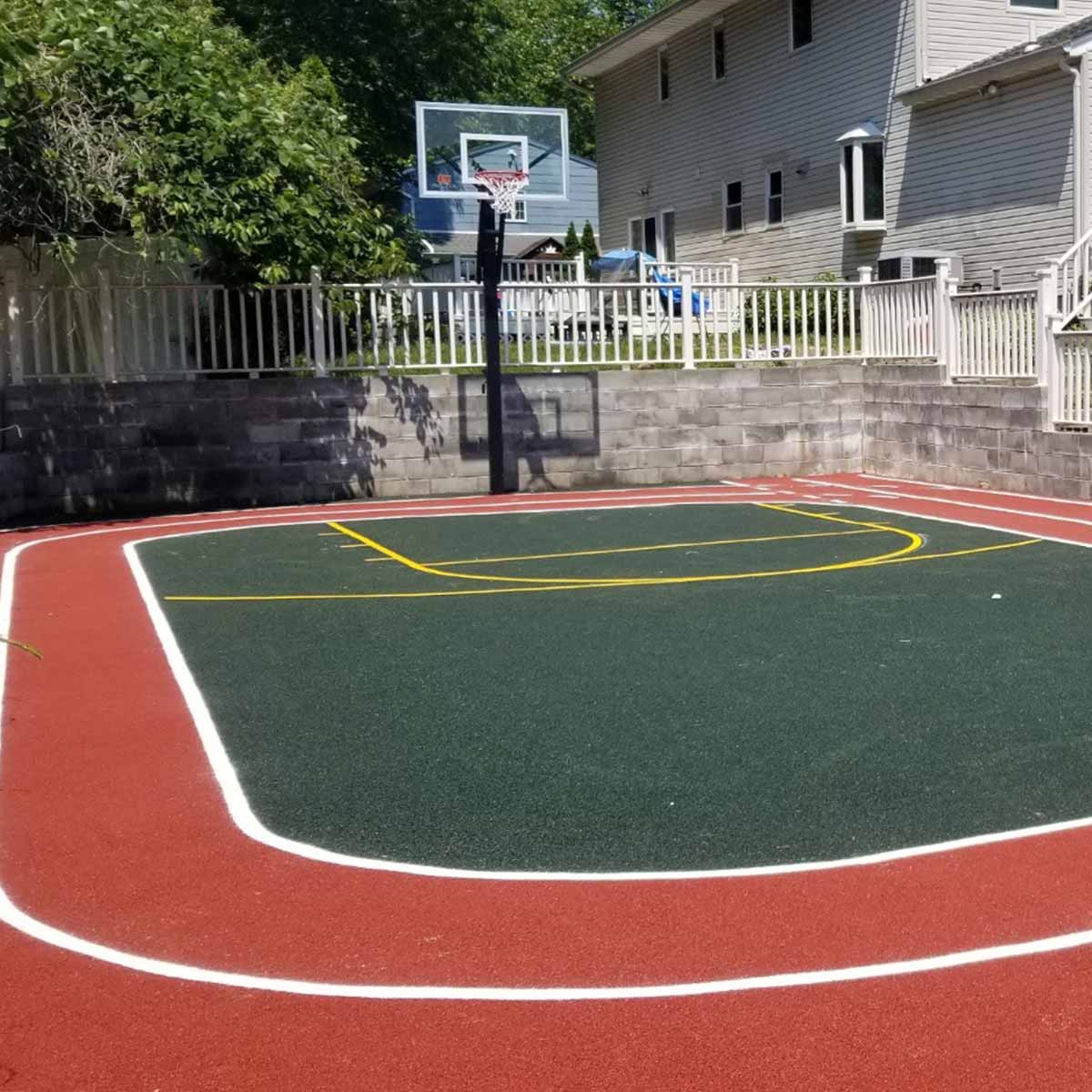 Outdoor basketball half-court with a red border, green playing surface, yellow and white lines, and a single hoop, located next to a house and surrounded by a fence and stone wall.