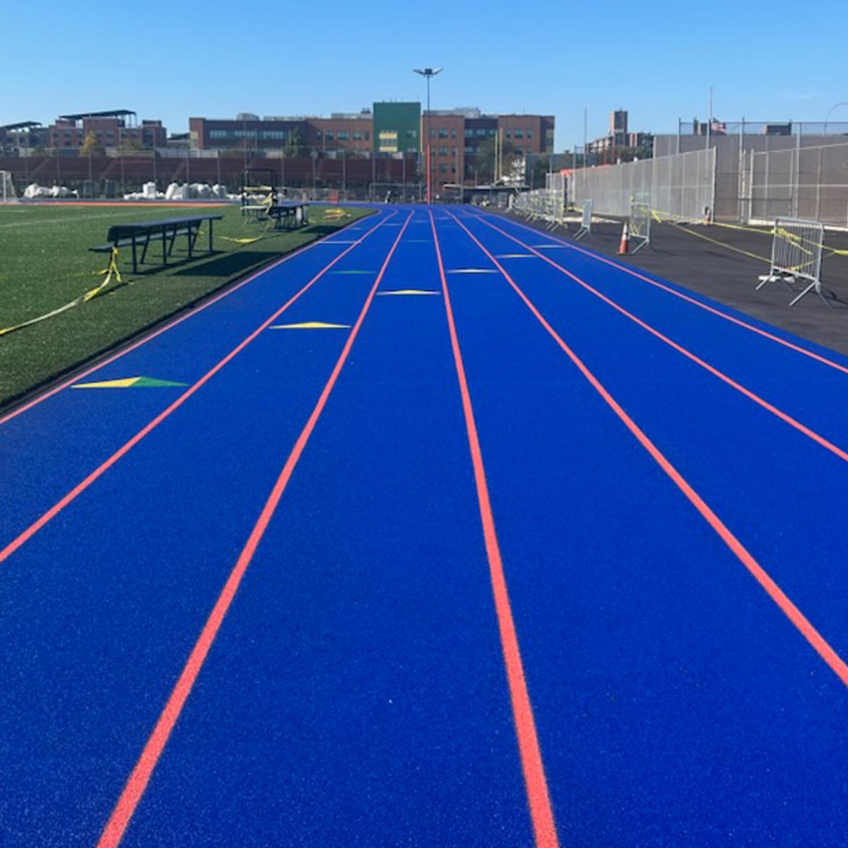 A bright blue running track with orange lane lines extends toward a cityscape under a clear sky, with a sports field and benches on the left and fencing on the right.