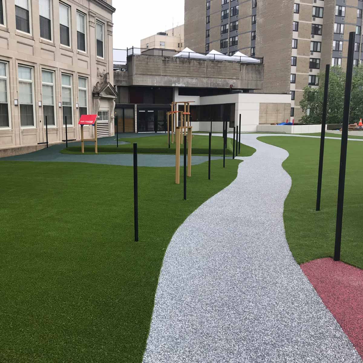 A winding gravel path runs through a grassy courtyard with young trees and black posts near a large building and apartment complex.