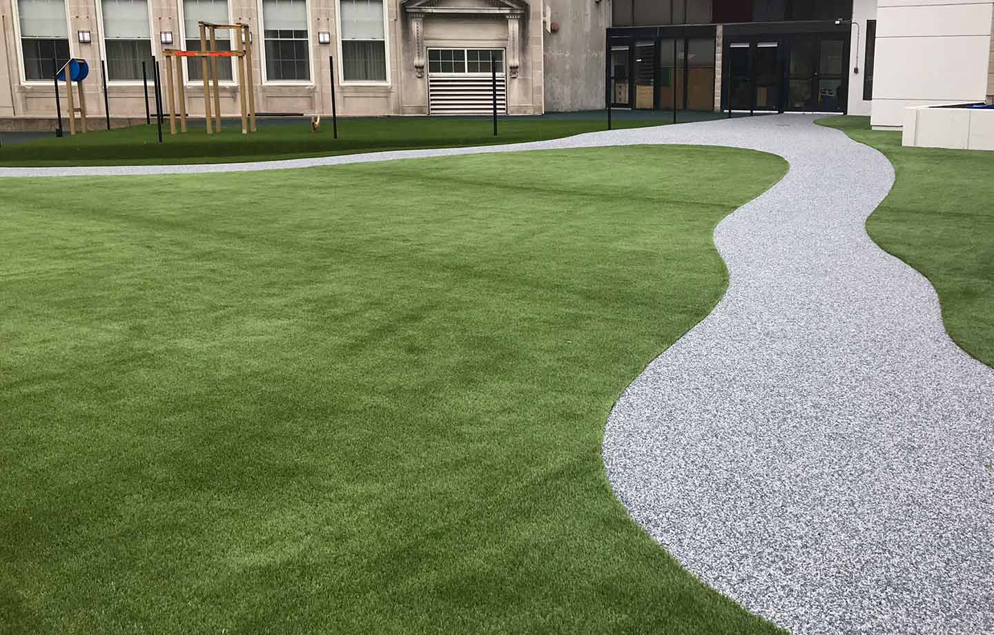 A winding gravel path runs through a neatly maintained artificial grass lawn near a modern building with large windows.