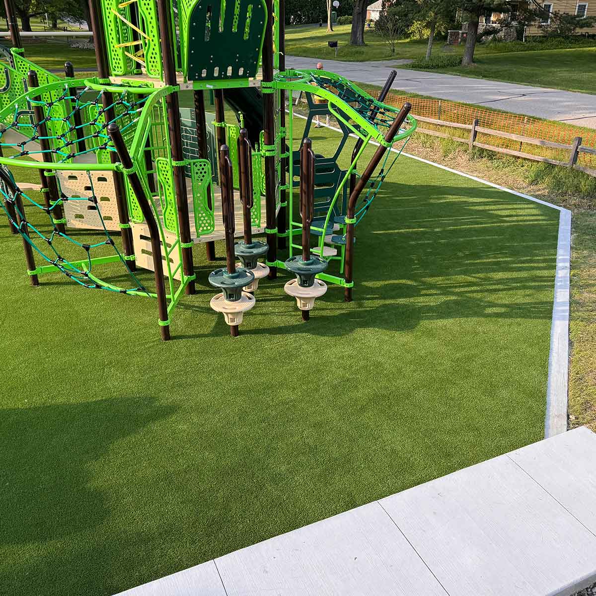 A modern playground structure with green and black climbing elements and spinning seats on artificial turf near a sidewalk and street.
