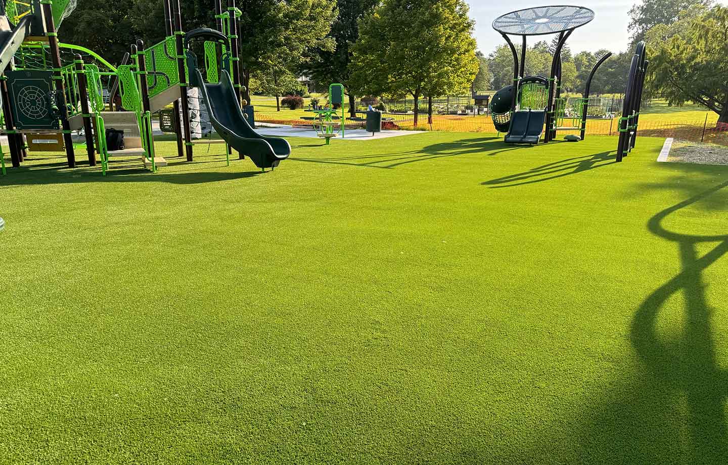 A playground with green artificial turf, slides, climbing structures, and swings, surrounded by trees and fencing under sunny skies.