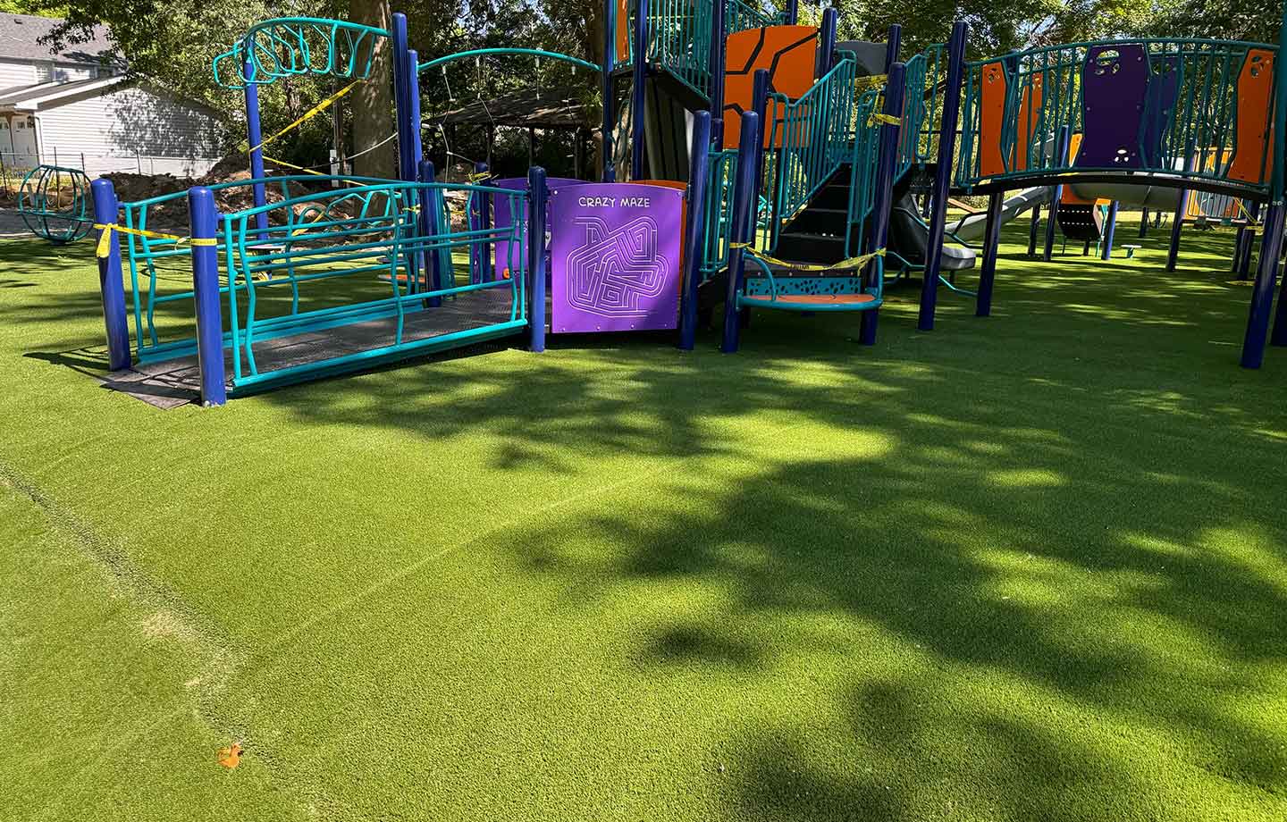 A playground with accessible equipment, including a ramp and maze panel, sits on artificial turf under the shade of trees.