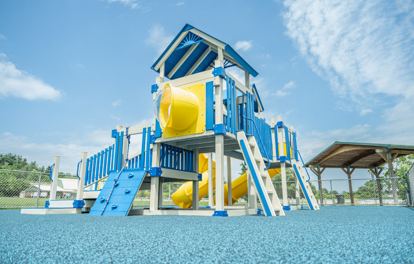Blue and yellow playground structure with slides and climbing features on a blue rubber surface, set outdoors under a partly cloudy sky.