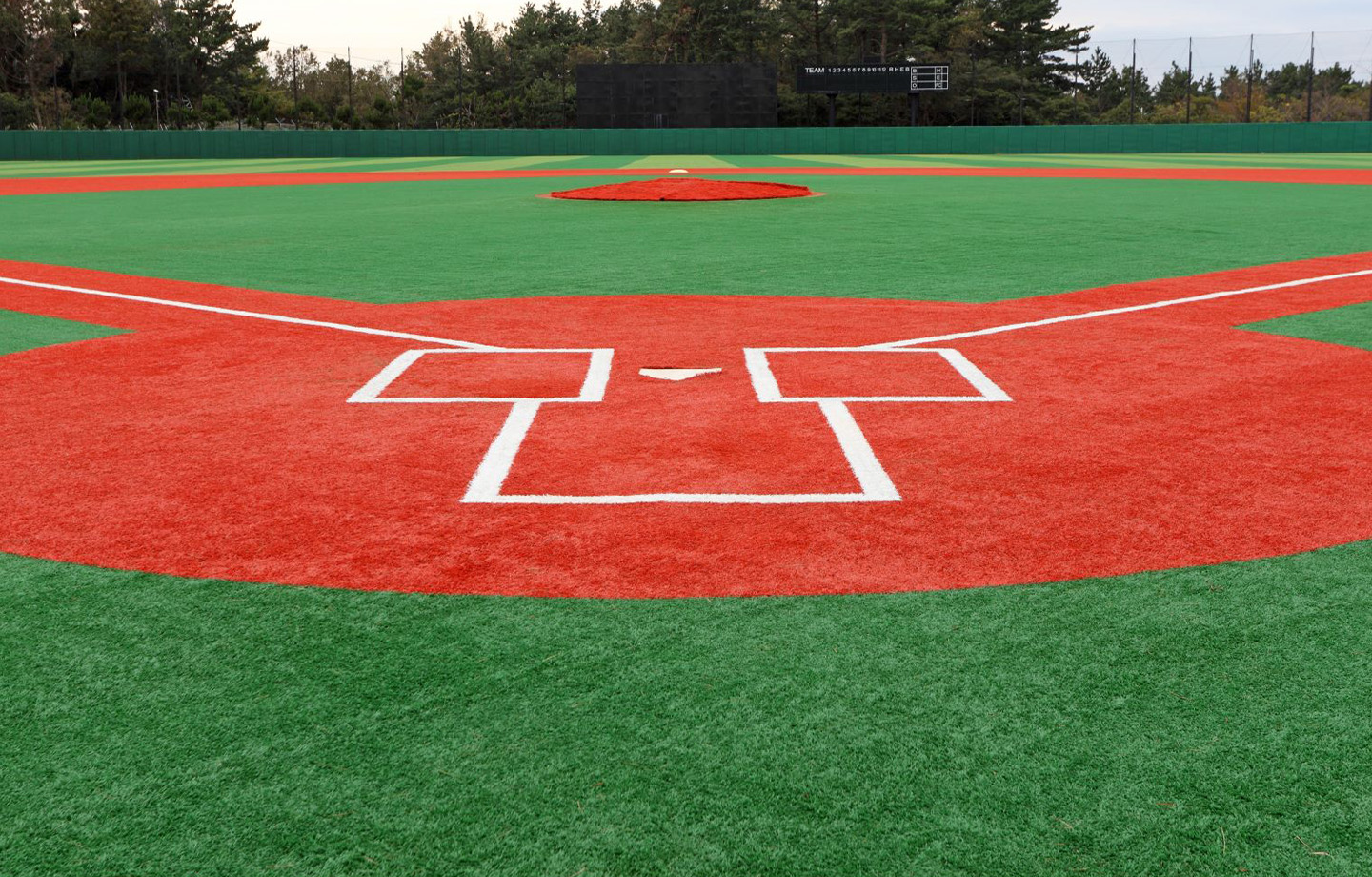 View of a baseball field’s home plate area with green artificial turf and red infield, featuring the base lines and a pitcher's mound in the distance.