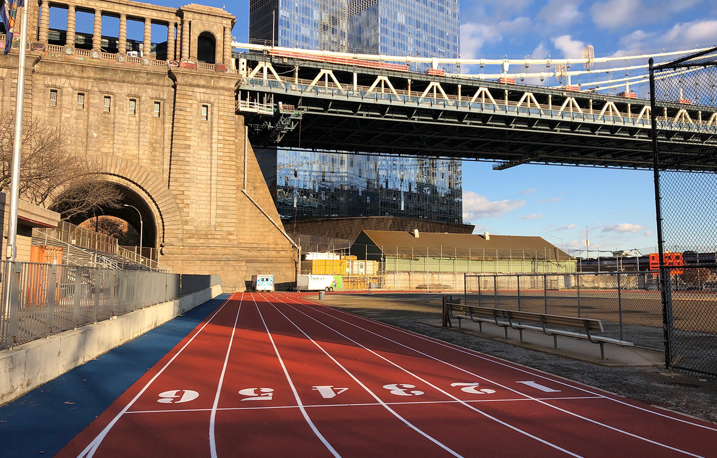 A red running track with six numbered lanes next to a baseball field, beneath a large bridge with tall buildings in the background.