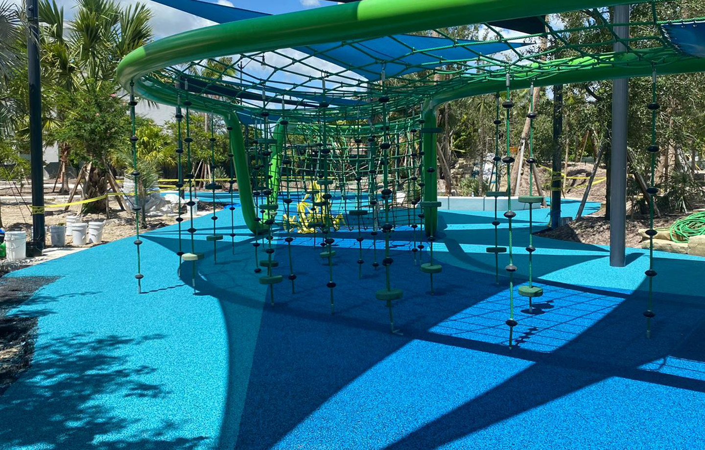 Outdoor playground with blue rubber flooring, green climbing structures, ropes, and platforms under a canopy, surrounded by trees and landscaping.
