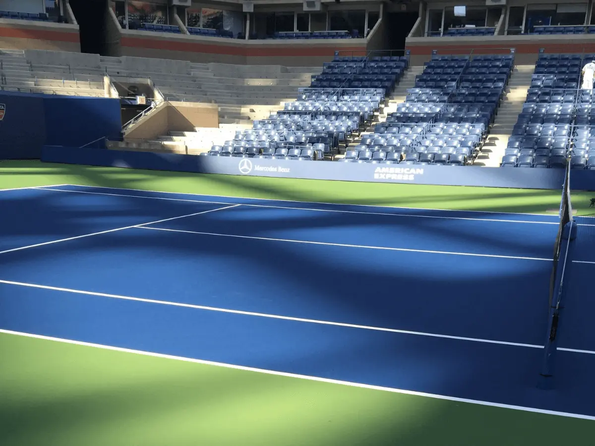 Empty tennis court with blue and green surface, net in the foreground, and rows of unoccupied blue seats in a stadium setting.